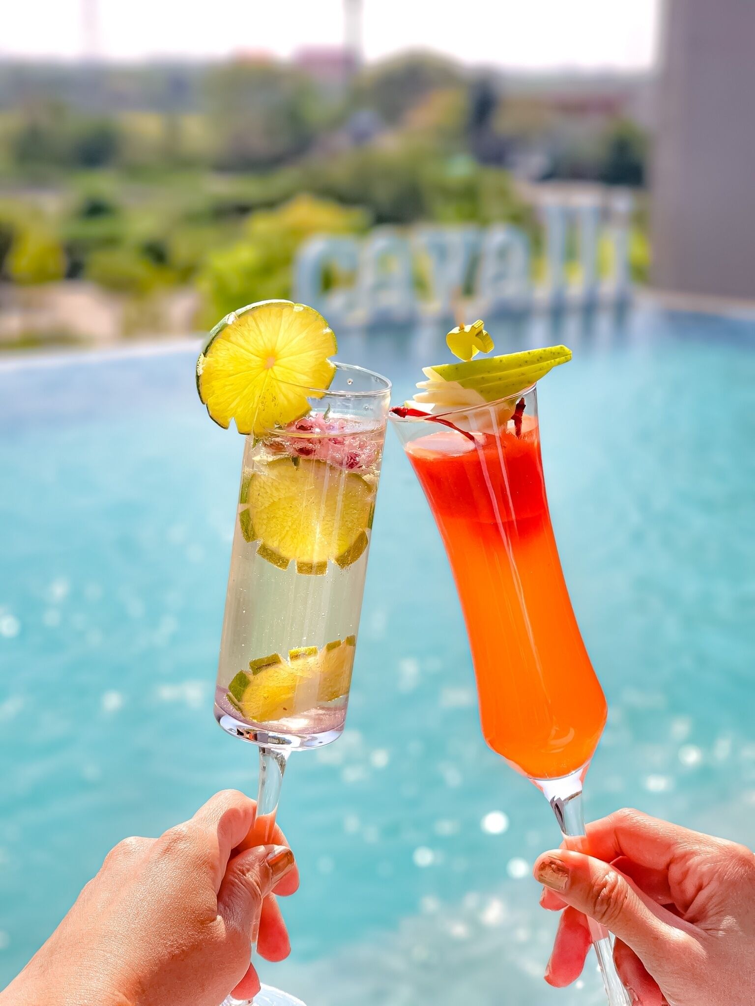 Deux cocktails, décorés de fruits ou d’autres éléments, tenus dans des mains, devant une piscine.