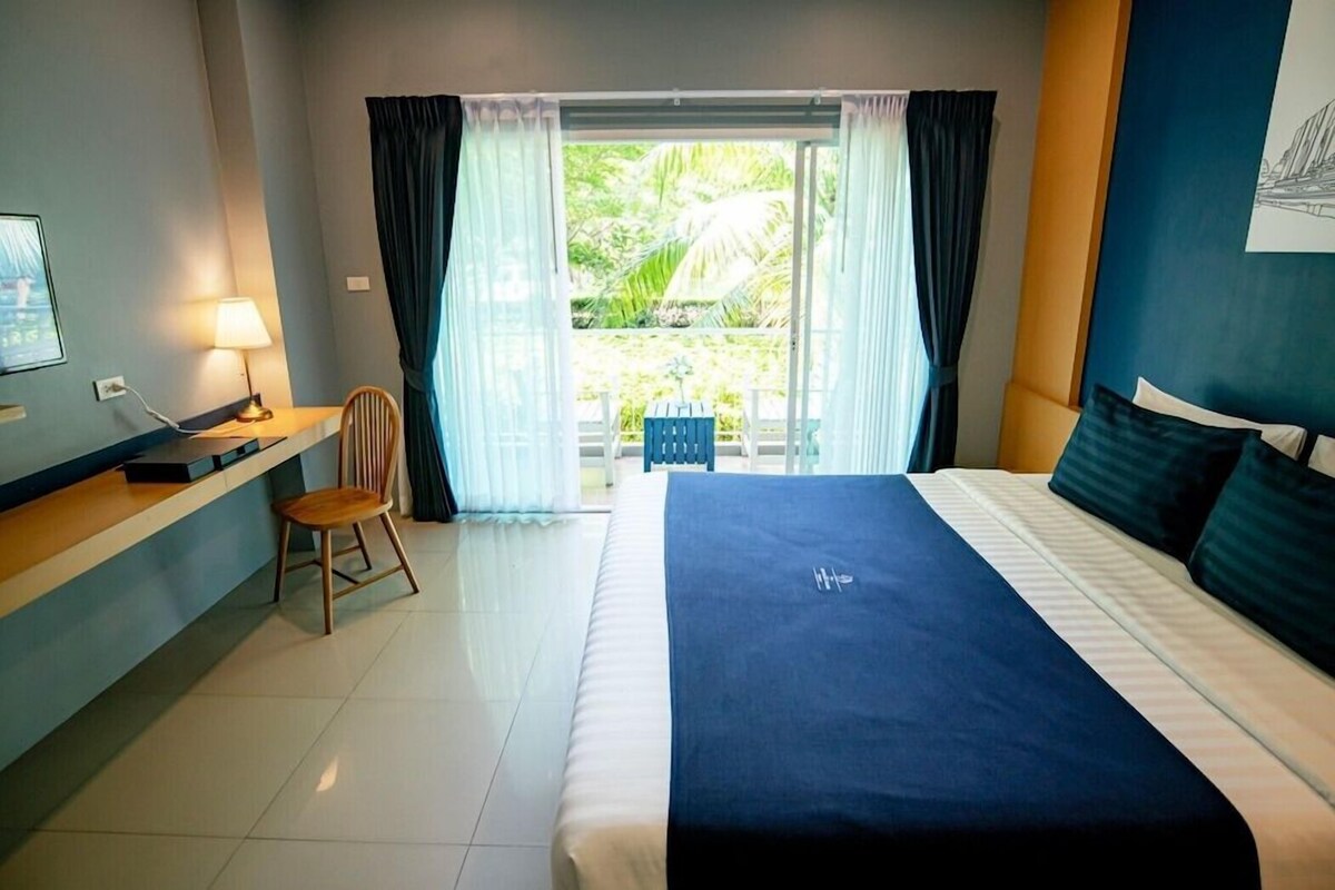 A hotel room with a bed, a desk, a chair, and a view of greenery through a window.