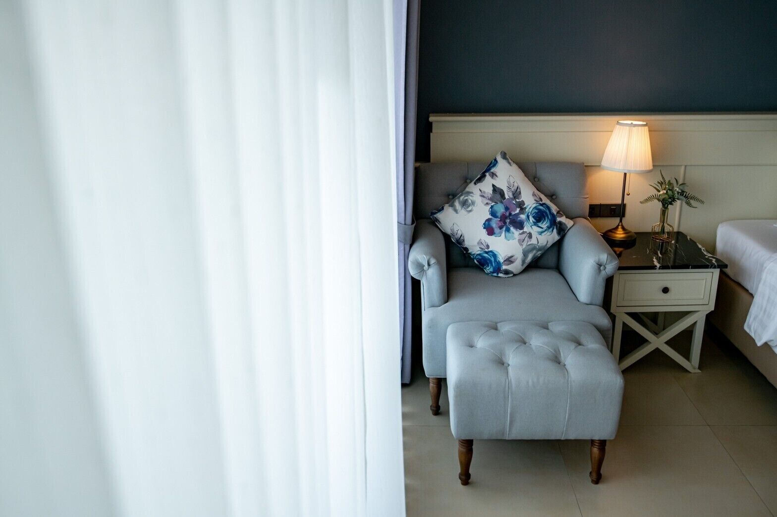 Una habitación de hotel con un sillón tapizado en gris, una mesita de noche con lámpara y un cojín estampado con motivos florales.