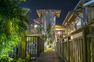 Uma passarela de madeira que leva a um edifício de design moderno, cercado por vegetação exuberante.