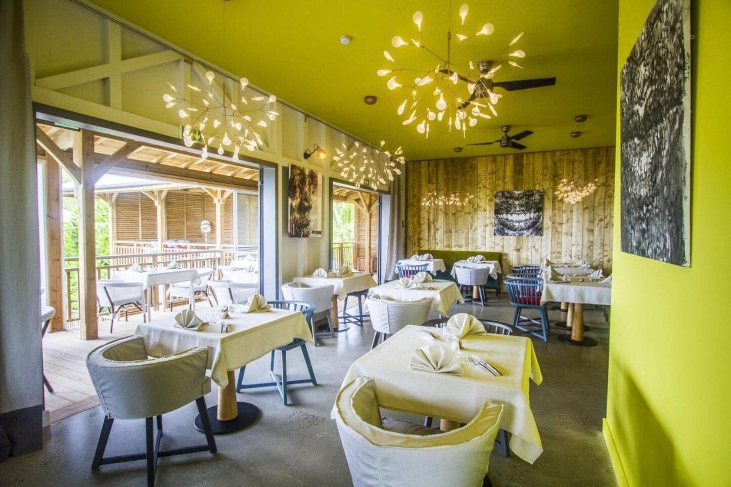 Een restaurant met een heldergeel interieur, houten wanden en moderne verlichtingsarmaturen. Tafels zijn gedekt met witte tafelkleden en stoelen.