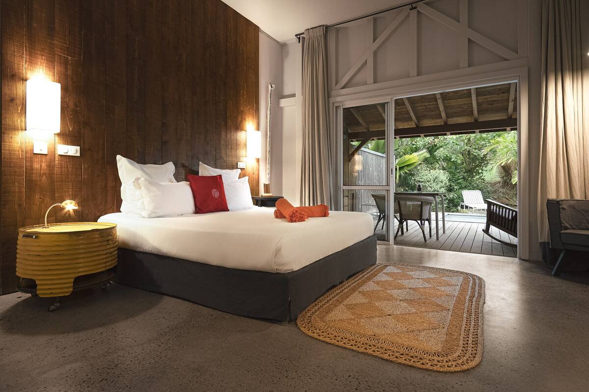 A bedroom with a large bed, a bedside table, a lamp, a woven rug, and a view of the outdoors.