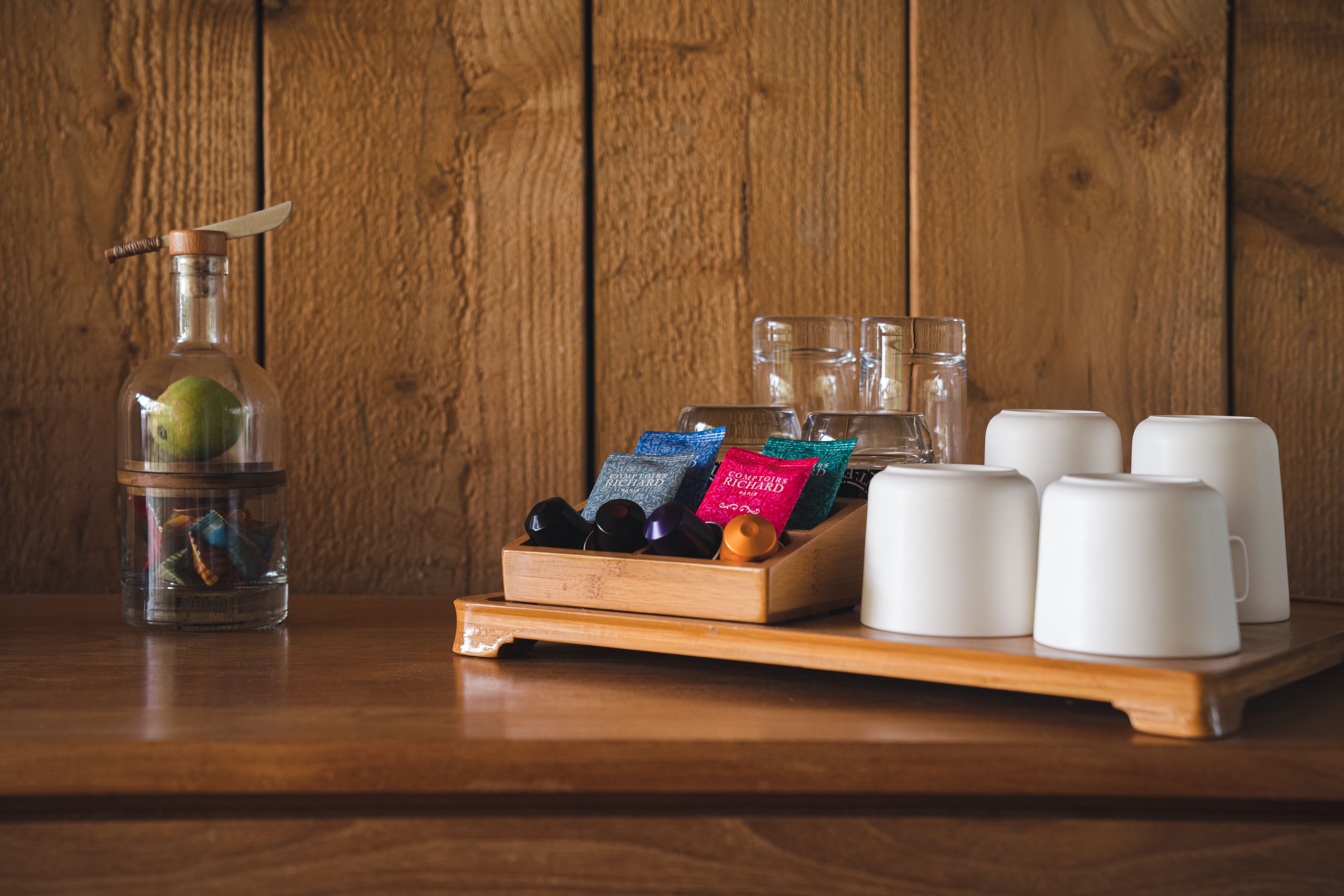 Una mesa de madera con una botella de vidrio, una bandeja de madera con tazas de café y paquetes, y una pared de madera.