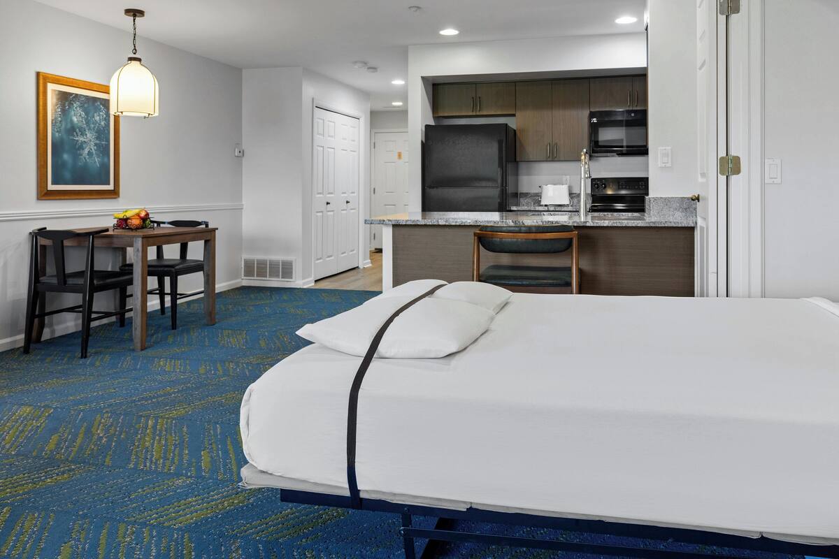 A hotel room with a bed, a desk, a chair, a kitchenette, and a painting on the wall.