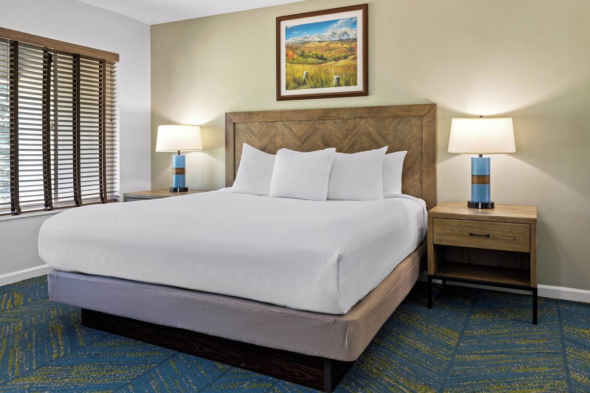 A hotel room with a large bed, two bedside tables with lamps, a wooden headboard, a painting on the wall, and a patterned carpet.