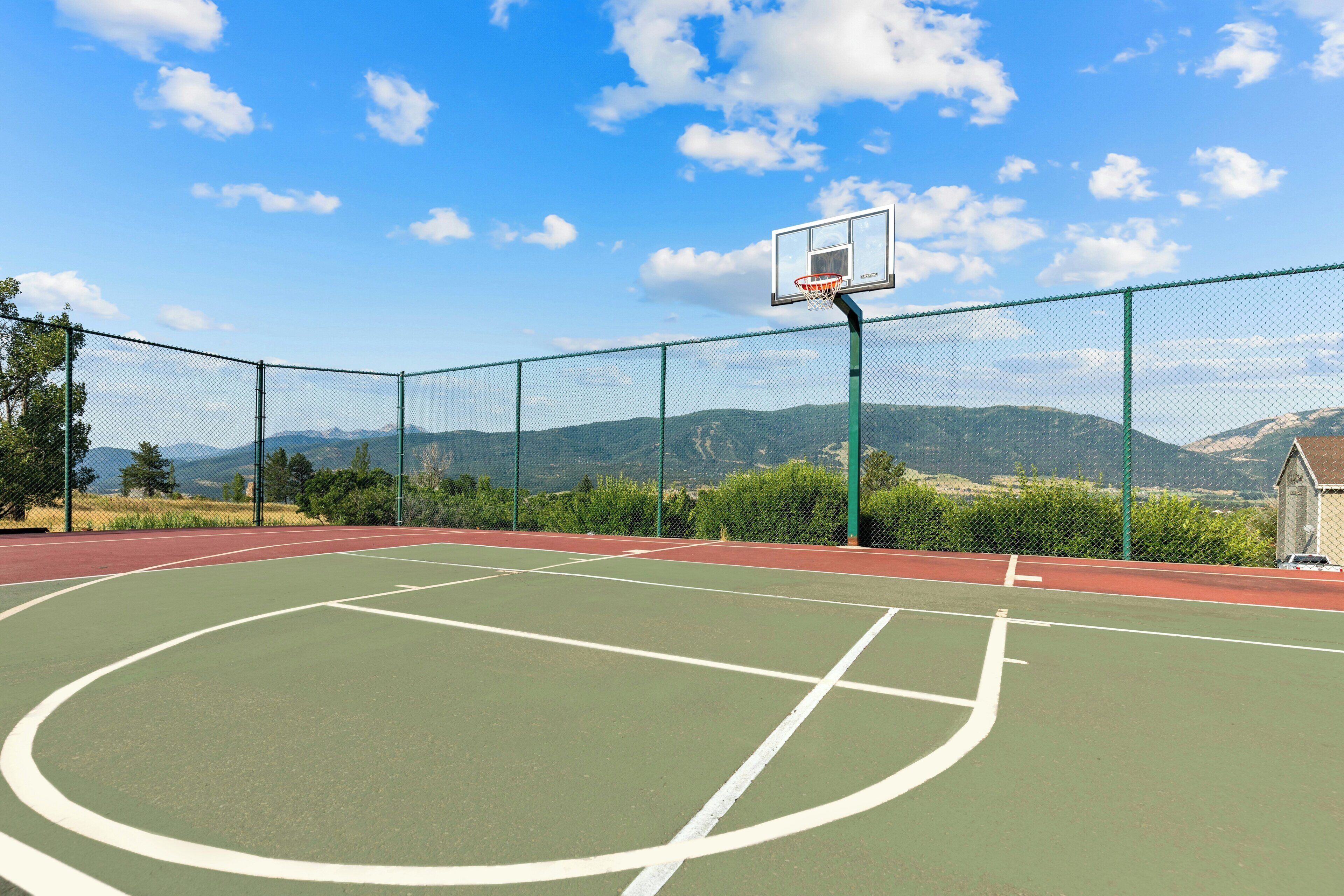 An outdoor basketball court with a chain-link fence, a basketball hoop, and a mountainous backdrop.