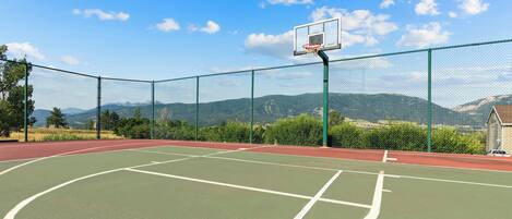 An outdoor basketball court with a chain-link fence, a basketball hoop, and a mountainous backdrop.