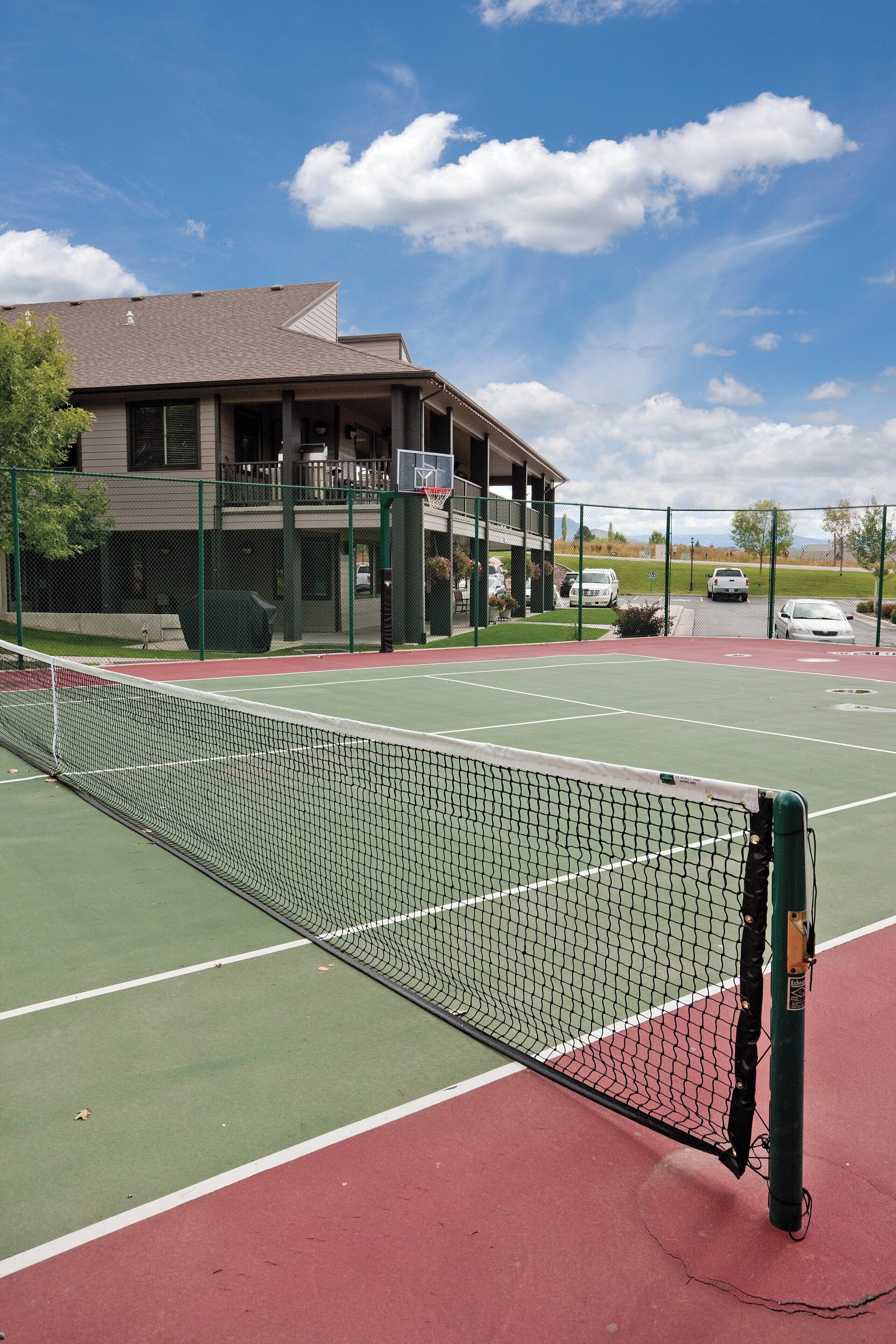 An outdoor tennis court with a net, green and red playing surfaces, and a building with a balcony in the background.