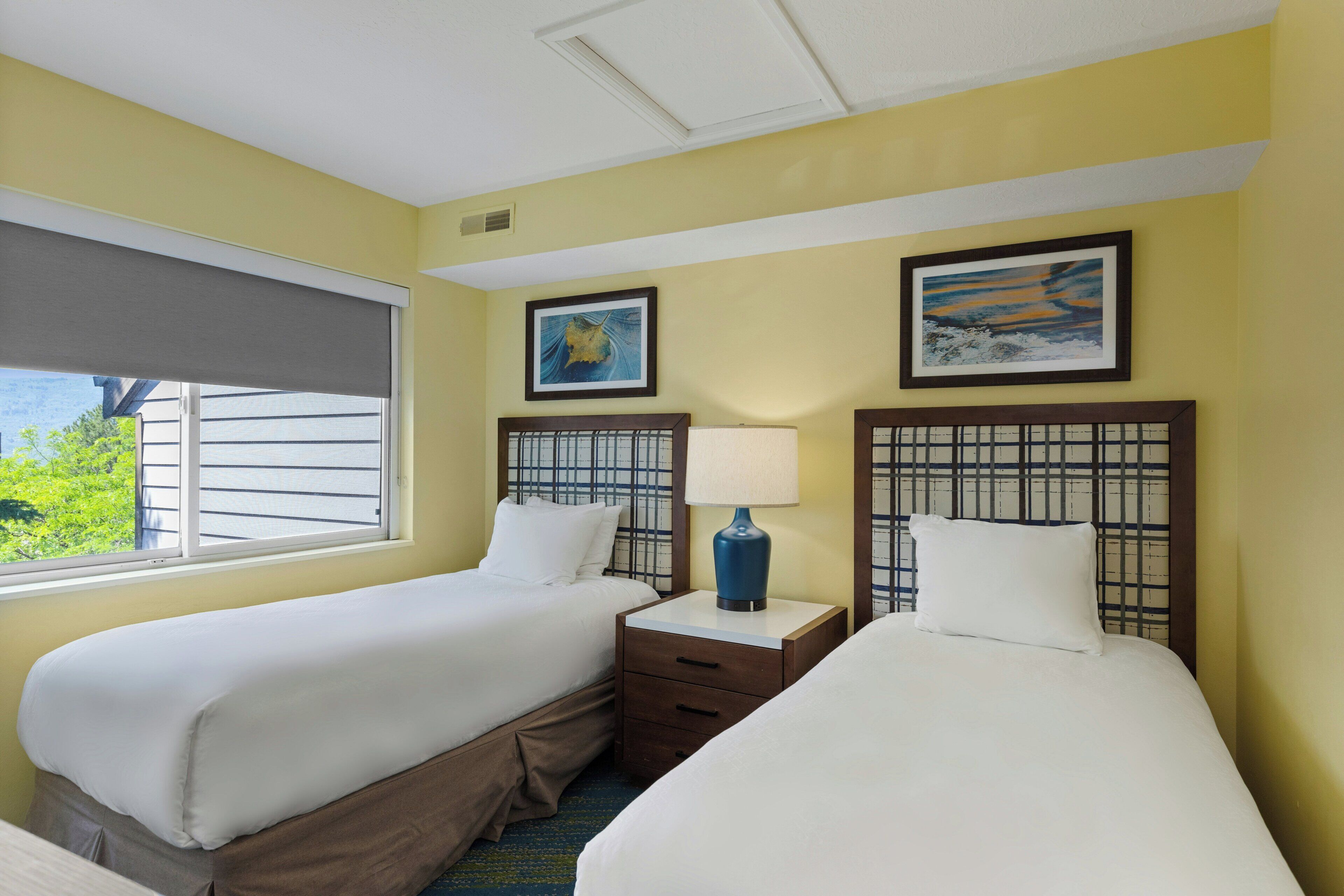 A hotel room with two beds, a window with blinds, a nightstand with a lamp, and artwork on the walls.