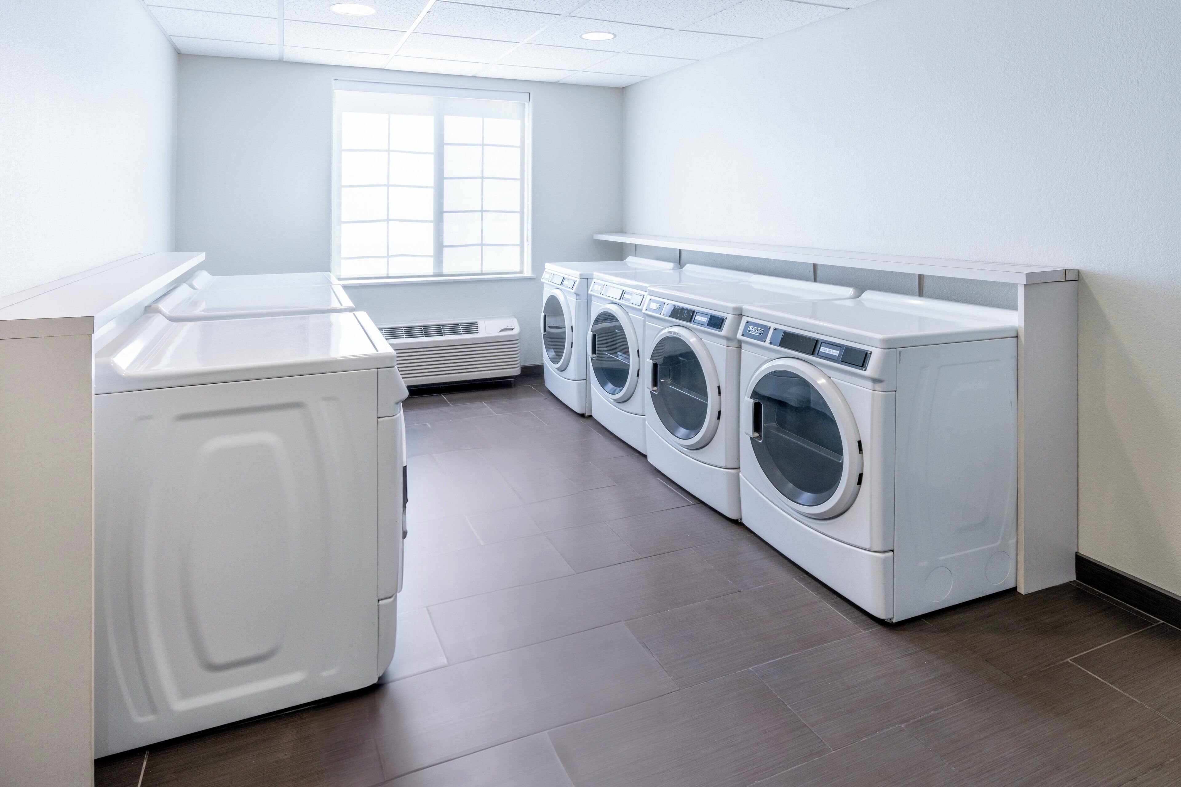 A laundry room with four front-loading washers and dryers, a large window, and a tiled floor.