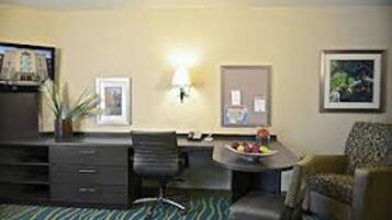 Une chambre d’hôtel avec un bureau, une chaise et une petite table sur laquelle se trouve des fruits. Il y a des lampes fixées au mur et des tableaux encadrés.