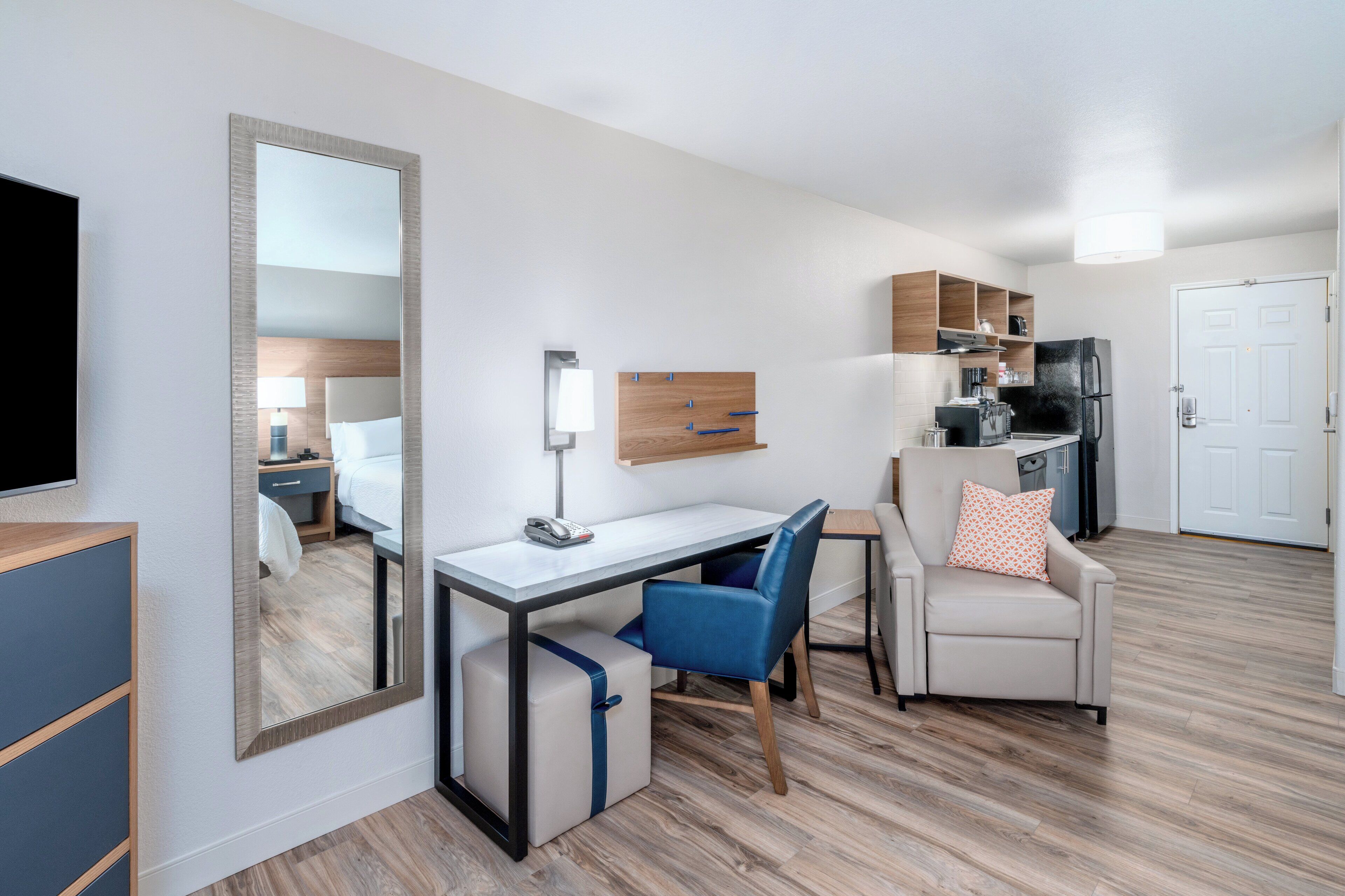 A compact hotel room with a bed, a desk, a chair, a TV, a mirror, a wardrobe, a kitchenette, and a door.