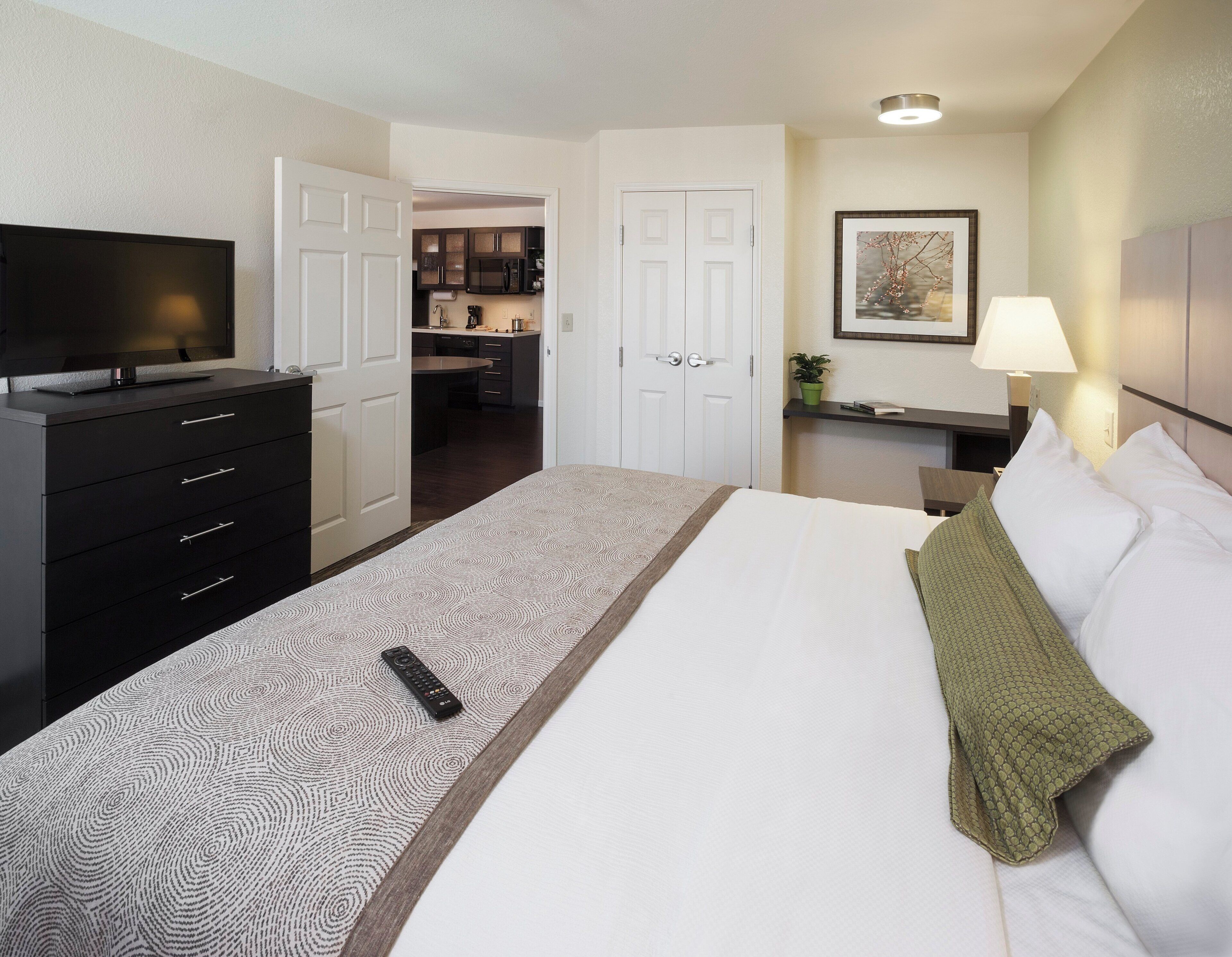 A hotel room with a bed, a TV on a dresser, a desk with a lamp, and a kitchen area visible through an open door.