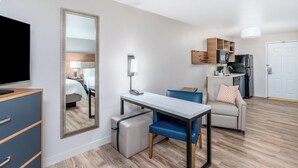 Une chambre d’hôtel moderne équipée d’une télévision, d’un bureau avec une chaise, d’un canapé, d’un coin cuisine et d’un grand miroir.