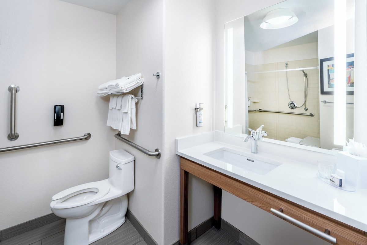 Een moderne badkamer met een wit toilet, een wastafel en een spiegel. Er zijn handgrepen aan de wanden en een handdoekrek met opgevouwen handdoeken.