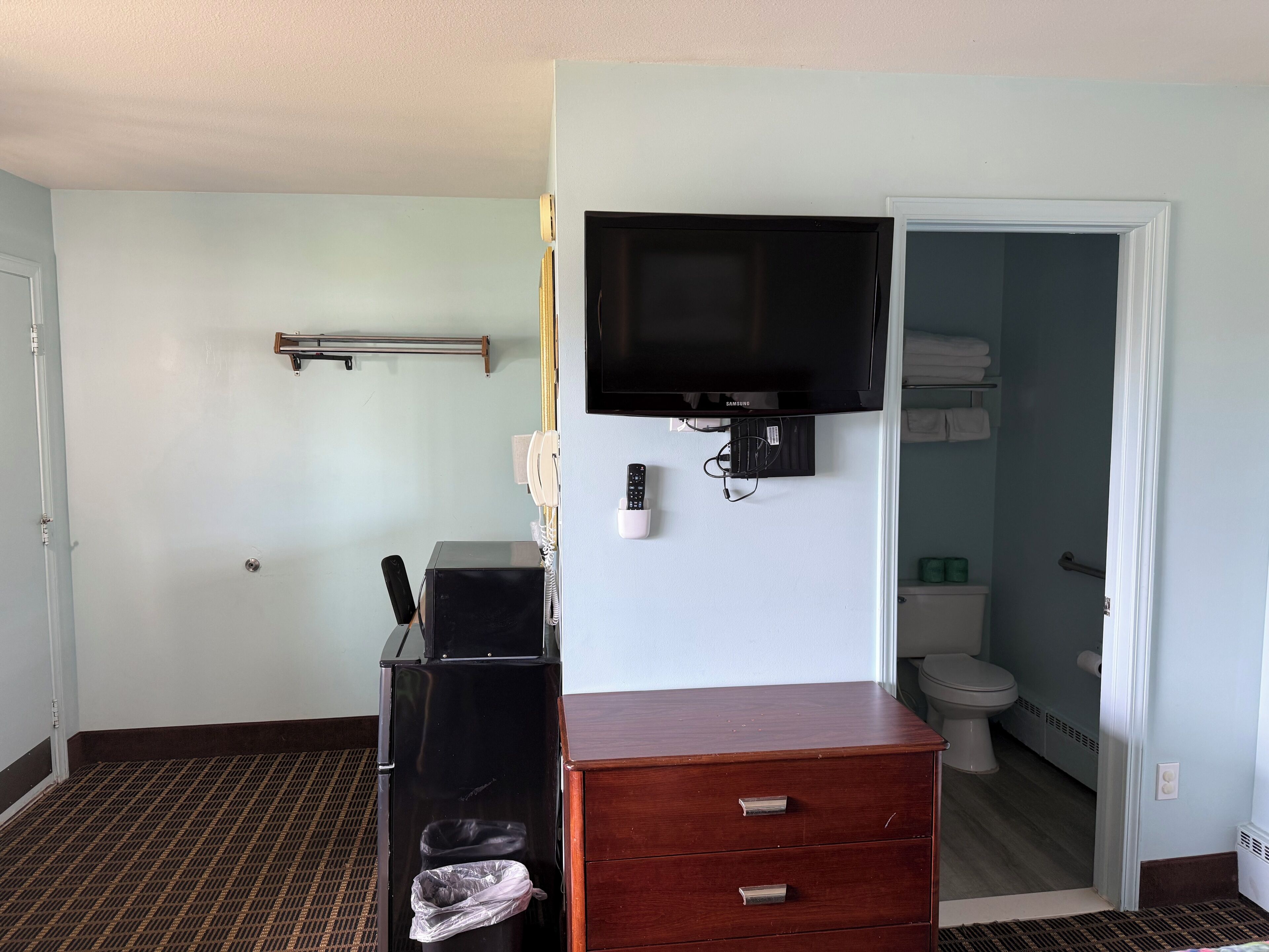 Une chambre d’hôtel équipée d’une télévision, d’un bureau avec une corbeille à papier, d’une armoire en bois, d’un téléphone et d’une salle de bain visible à travers une porte ouverte.
