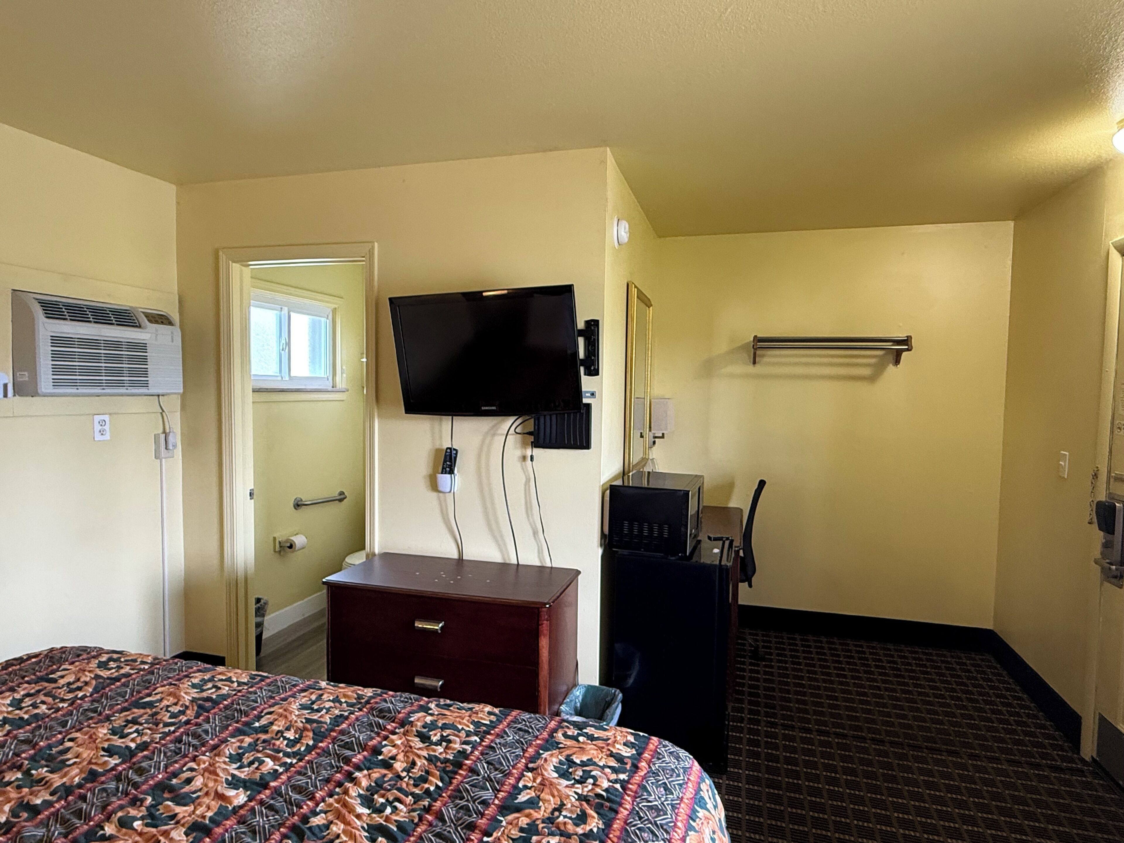 Una habitación de hotel con cama, cómoda, televisor montado en la pared, espejo y calefactor.