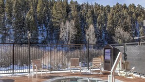 Ein Whirlpool auf einer Holzterrasse mit Stühlen und einem Handtuch, umgeben von einem Metallzaun und bewaldeter Landschaft.