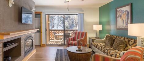 A cozy living room with a fireplace, a ceiling fan, a dining area with a view of snow-covered trees, and a large window.