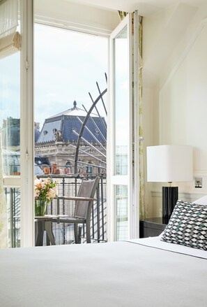 Habitación de hotel con balcón, una cama, una mesa con un jarrón de flores y una lámpara.