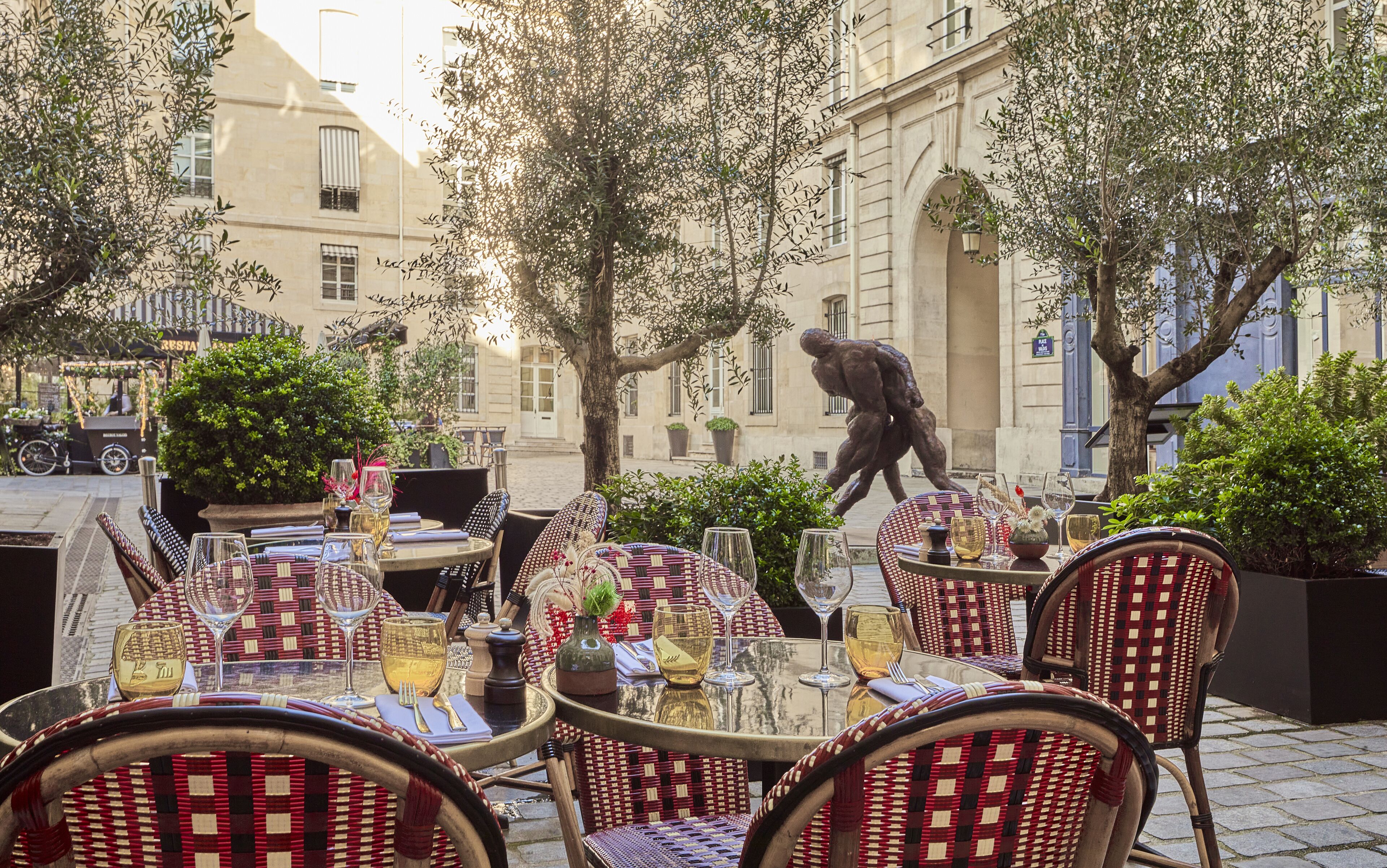 Un área de comedor al aire libre con mesas redondas y sillas de mimbre, rodeada de follaje y un edificio de arquitectura clásica.