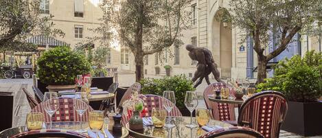 Un área de comedor al aire libre con mesas redondas y sillas de mimbre, rodeada de follaje y un edificio de arquitectura clásica.