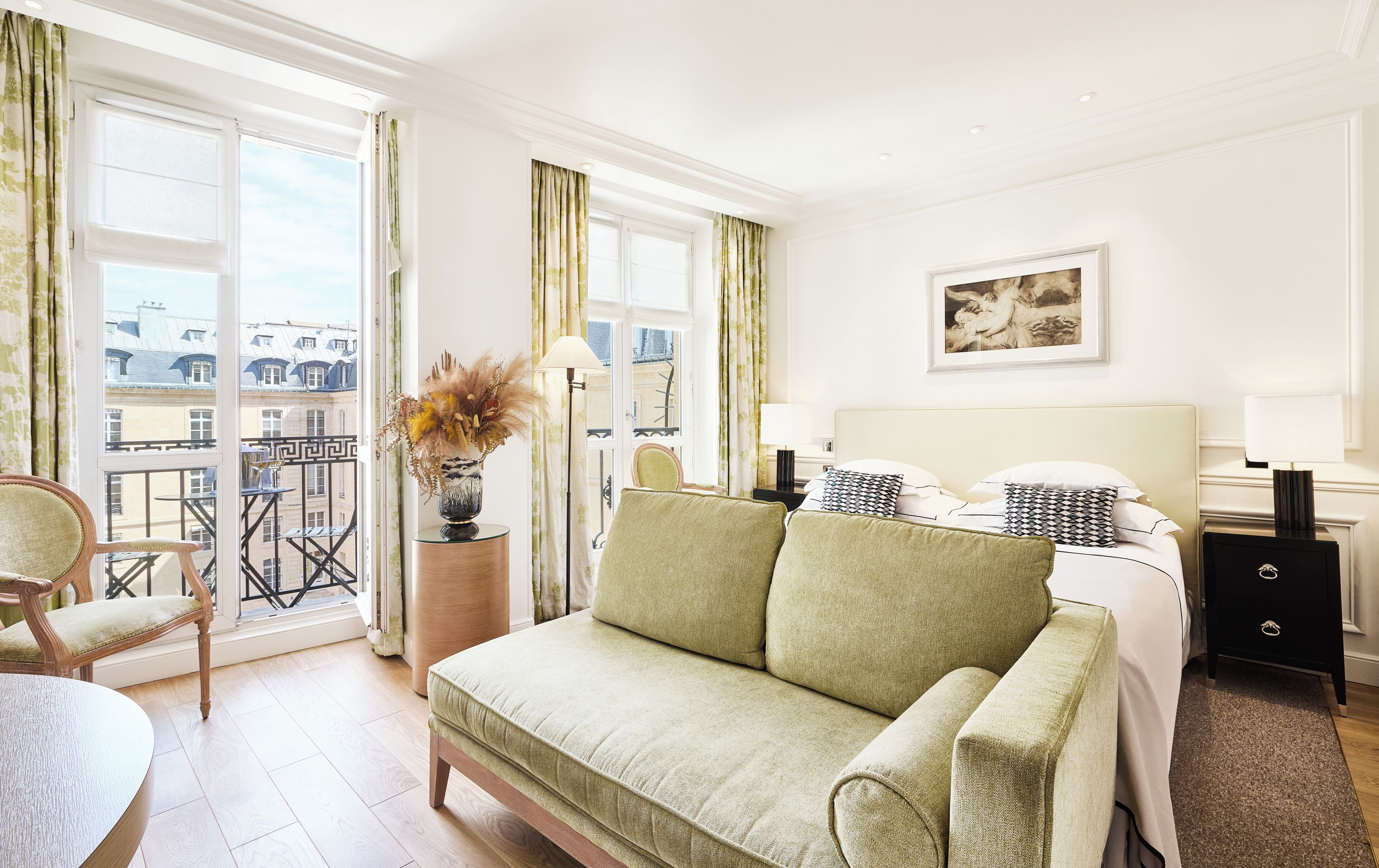Habitación de hotel con sofá verde, cama con almohadas estampadas, mesita de noche con un jarrón y balcón con vistas a edificios.