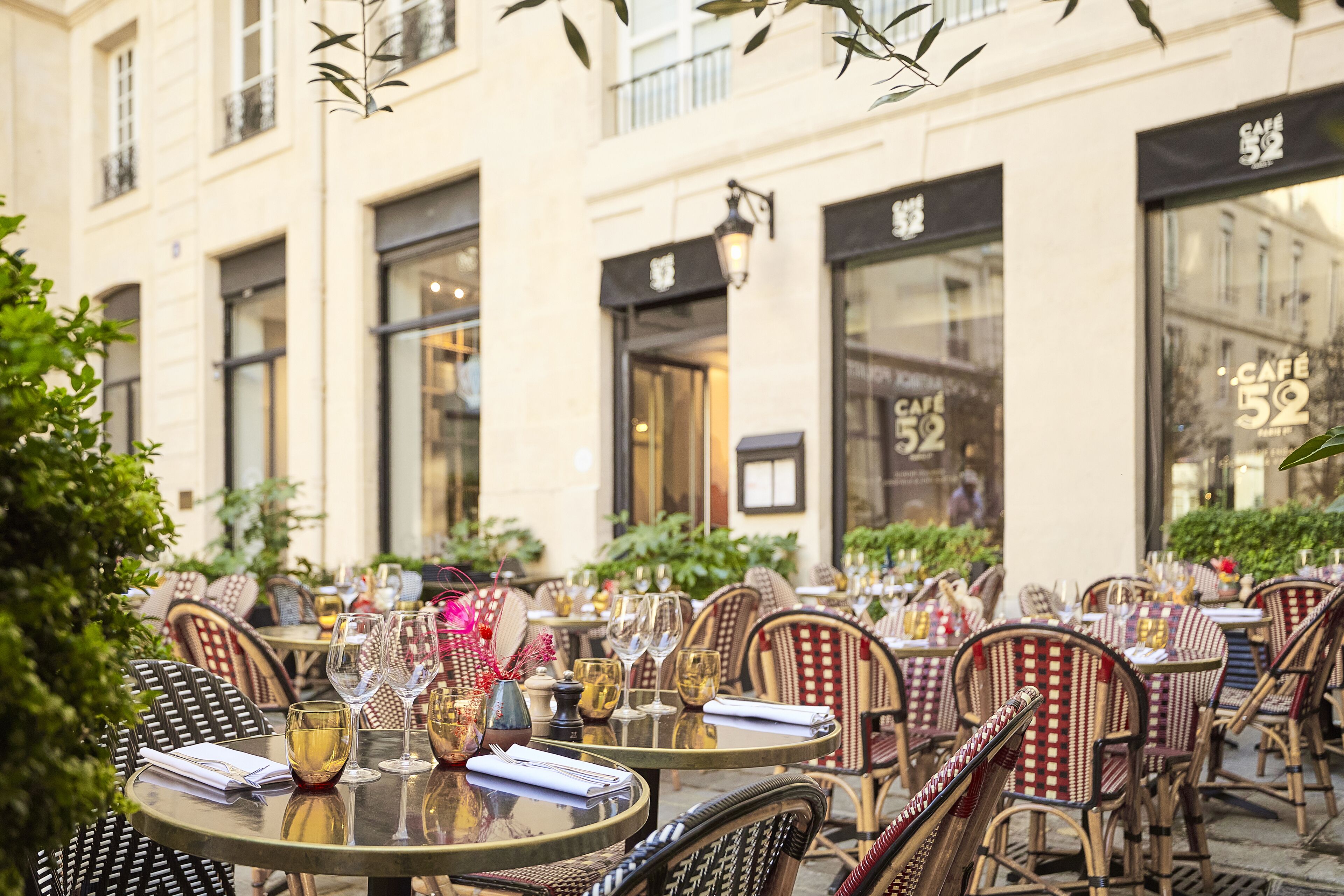 An outdoor café with round tables and wicker chairs, set against a building with large windows and a café sign.