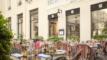 An outdoor café with round tables and wicker chairs, set against a building with large windows and a café sign.