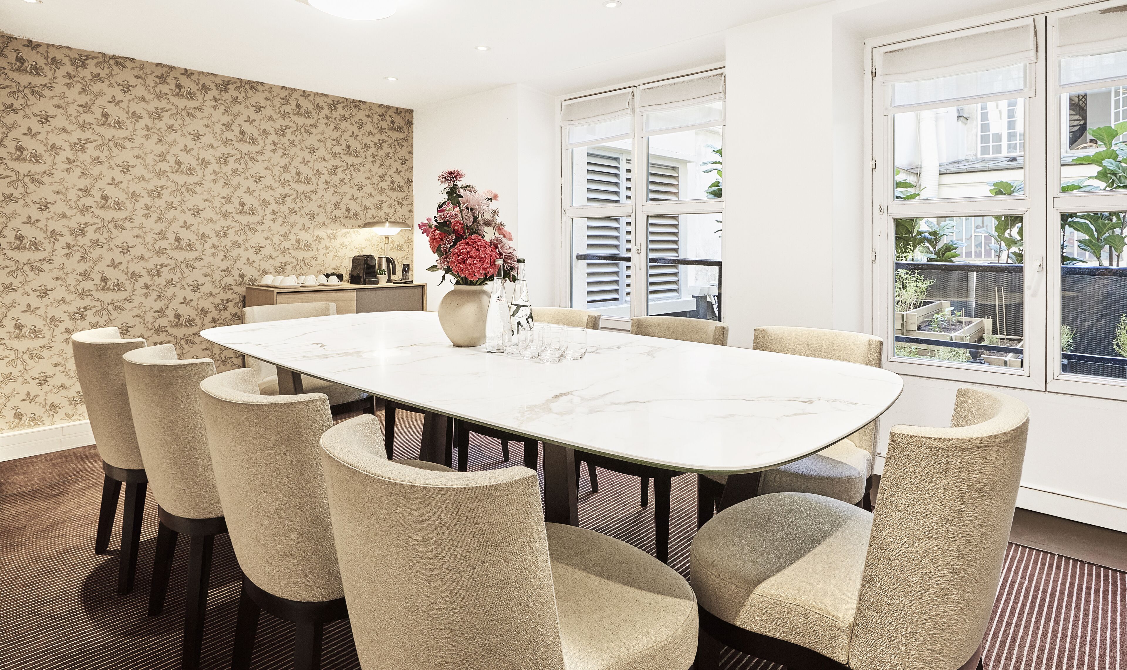 A dining area with a large table, six chairs, a vase with flowers, a coffee maker, and a lamp.