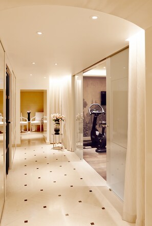 A modern hotel corridor with a glass wall, a fitness machine, and a seating area with chairs and a vase of flowers.