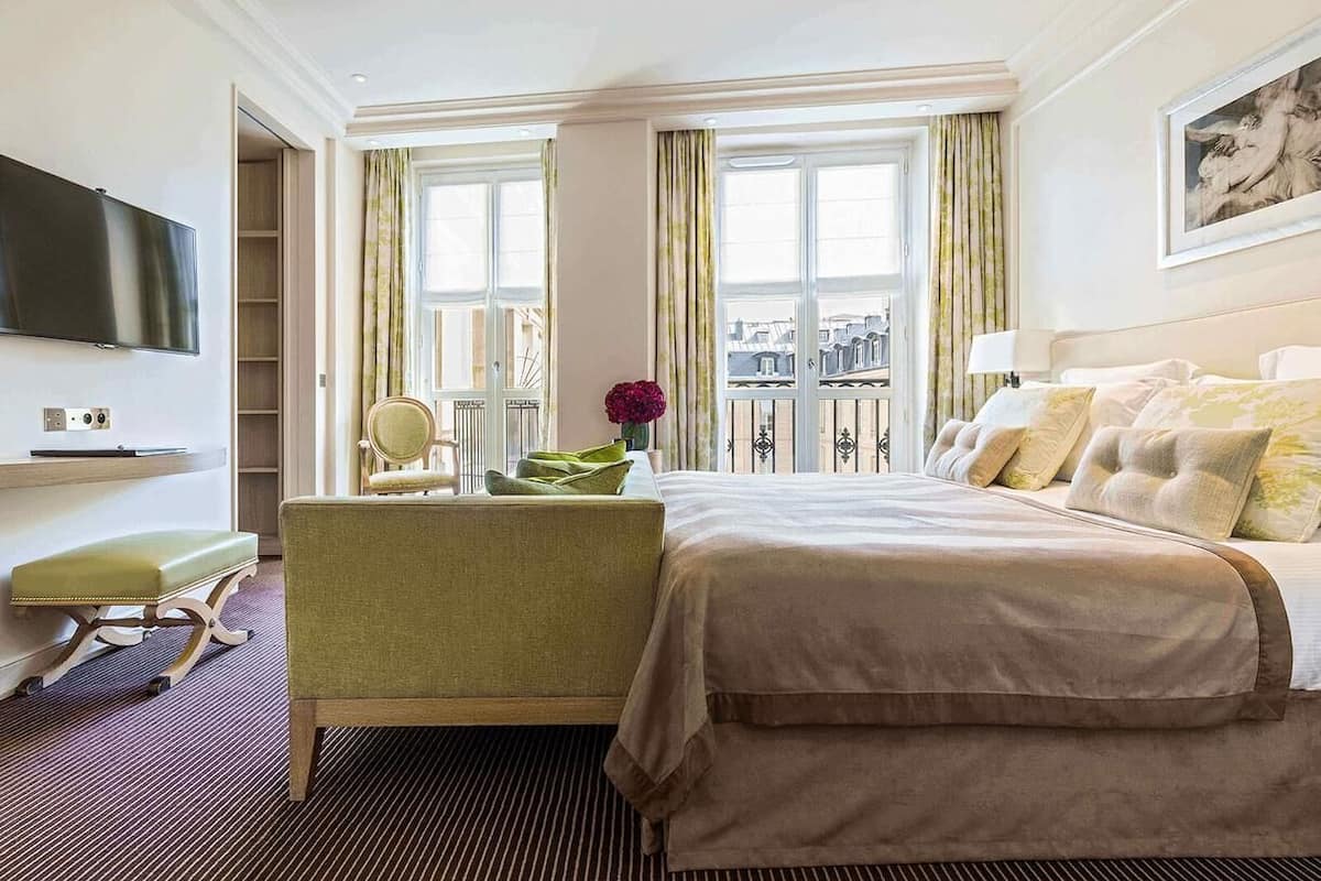 A hotel room with a bed, a chair, a TV, a window with curtains, and a balcony view.