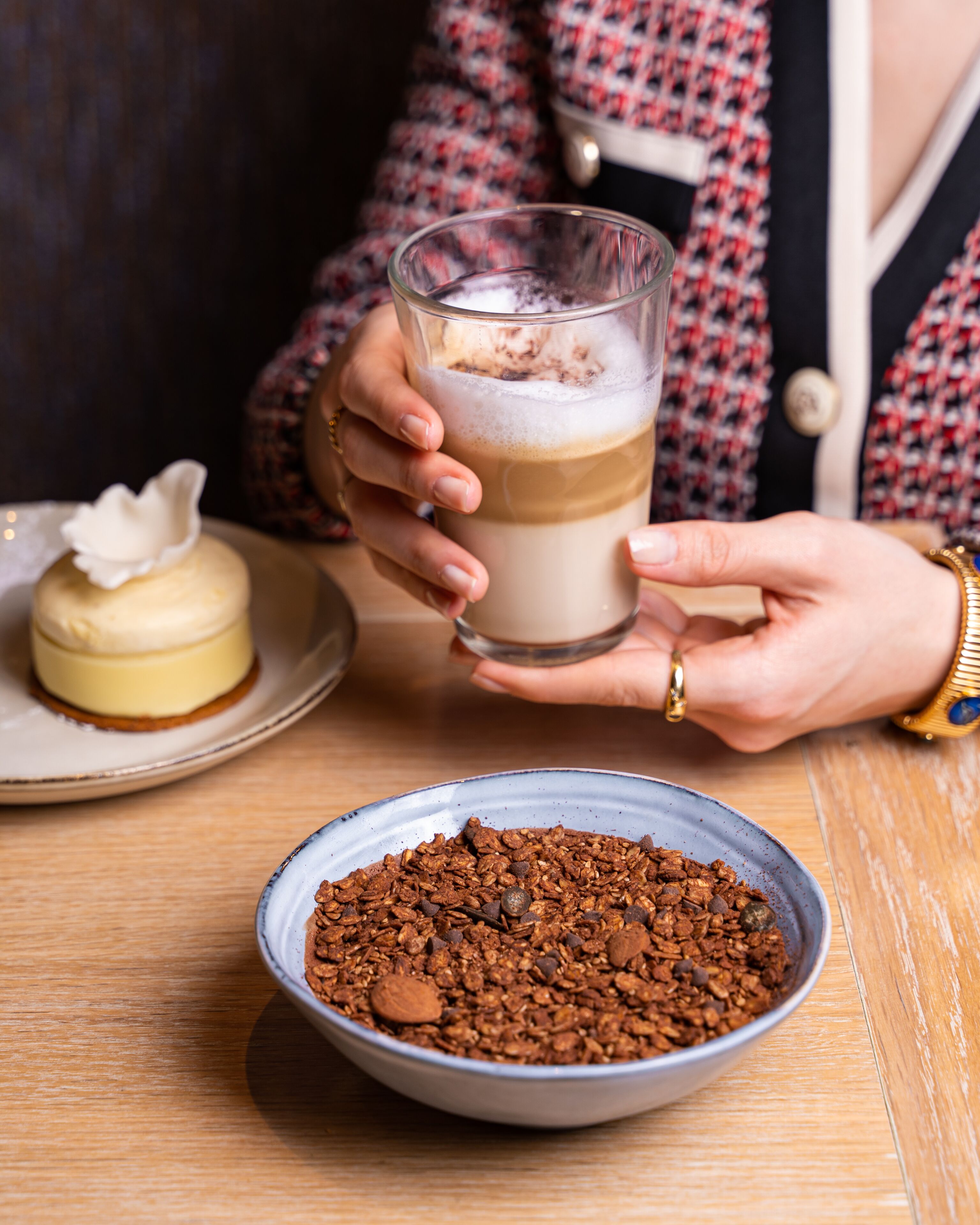 A person holding a glass of layered beverage, a bowl of granola, and a dessert on a table.
