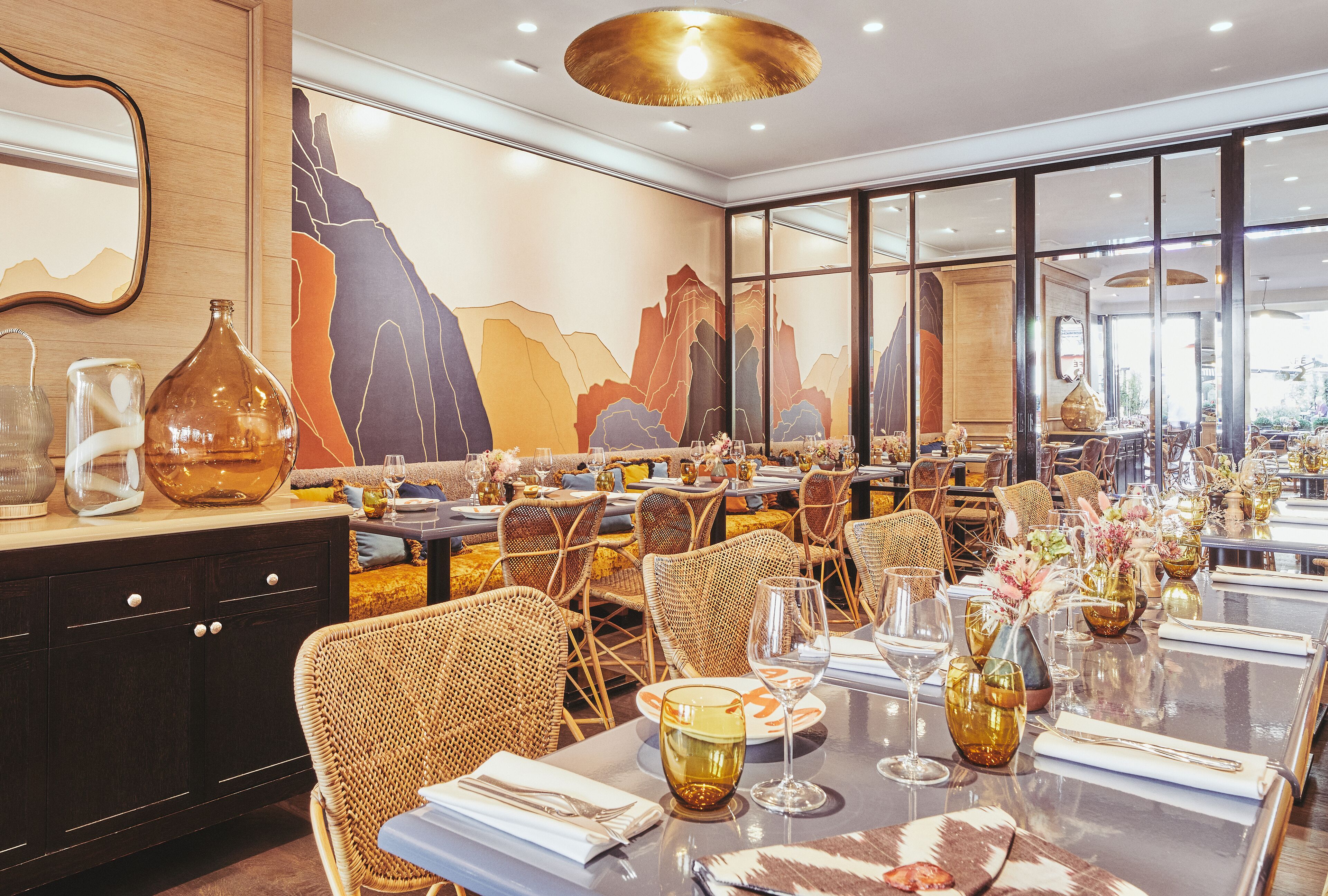 A restaurant with a long dining table set for multiple guests, wicker chairs, and a large mural on the wall.