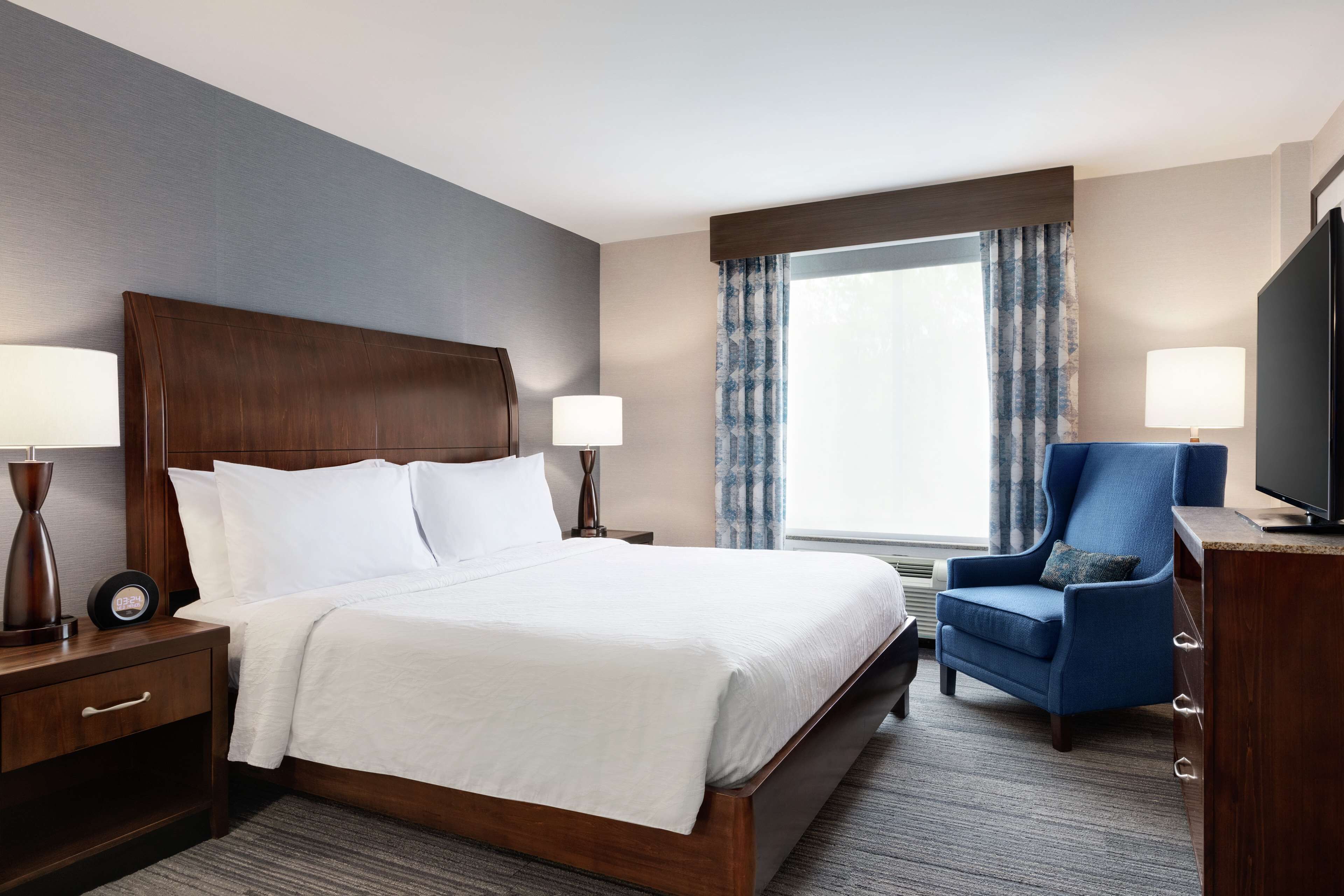 Ein Hotelzimmer mit einem großen Bett, zwei Nachttischen mit Lampen, einem Fernseher, einem blauen Sessel und einem Fenster mit Vorhängen.
