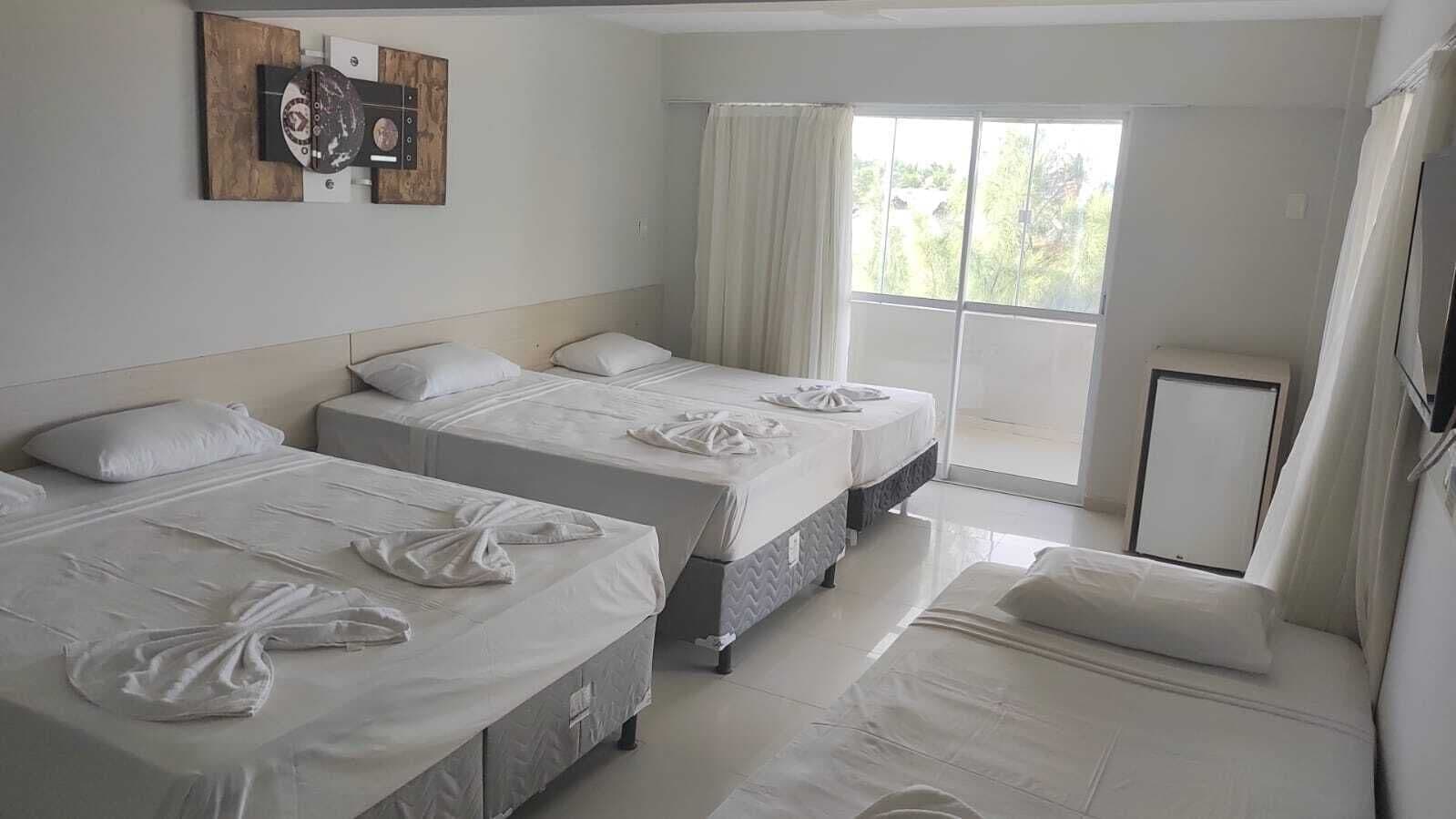 A hotel room with three beds, a large window with curtains, and a flat-screen TV on the wall.