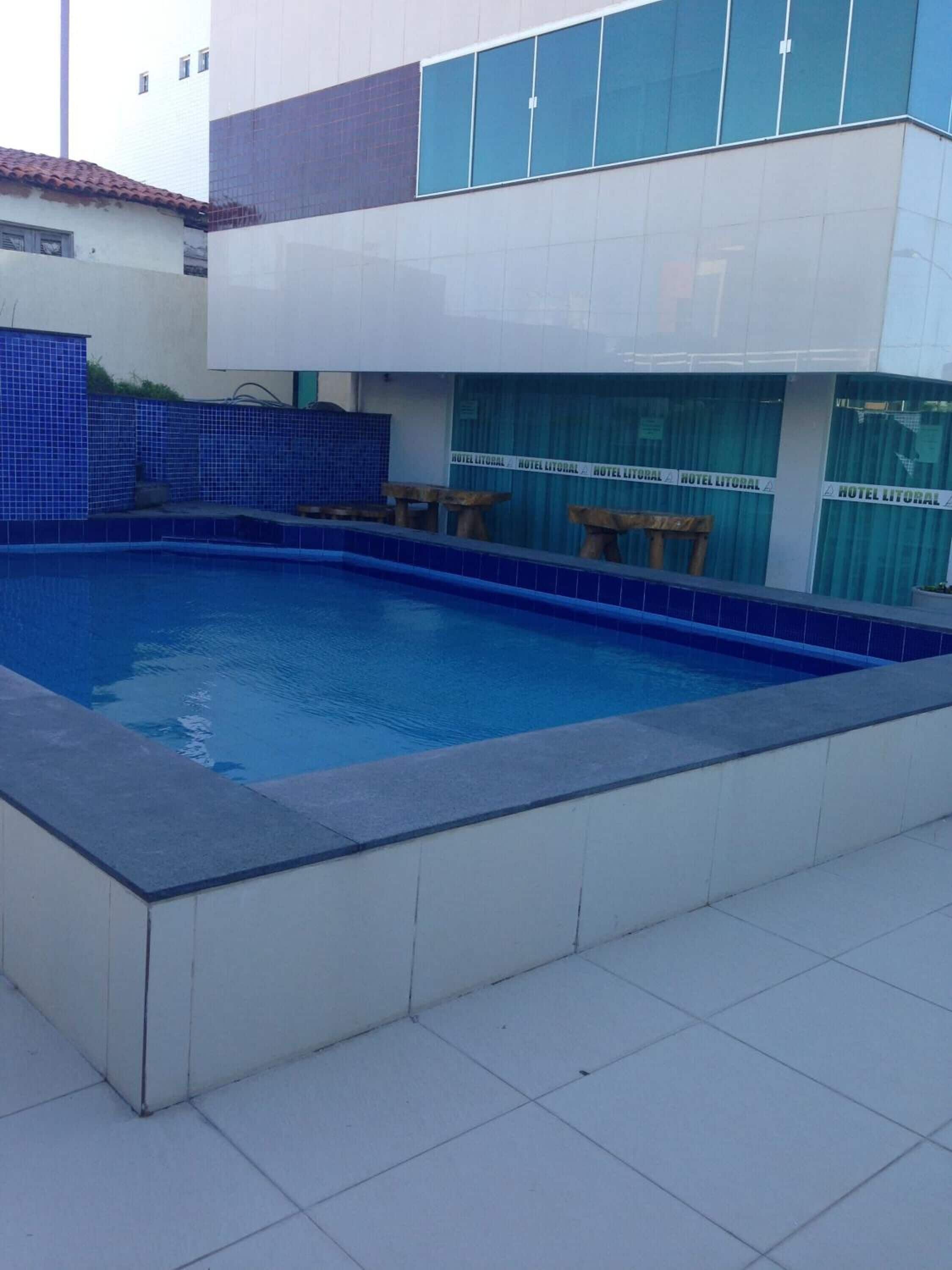 A swimming pool with a tiled deck, a covered seating area, and a building with large windows and a tiled facade.