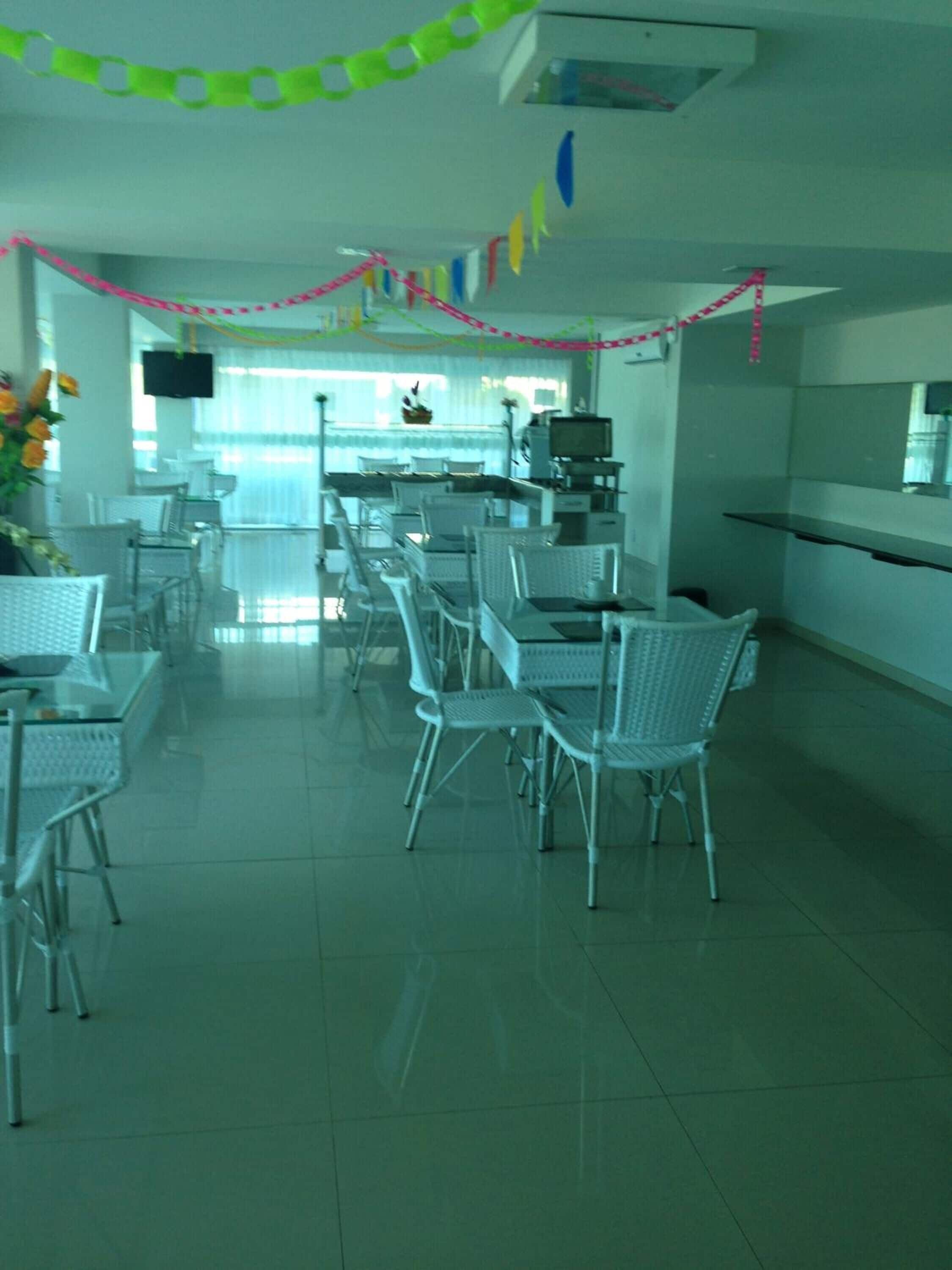 Un comedor amplio con sillas y mesas blancas, una cocina con mostrador y electrodomésticos, y un televisor montado en la pared.