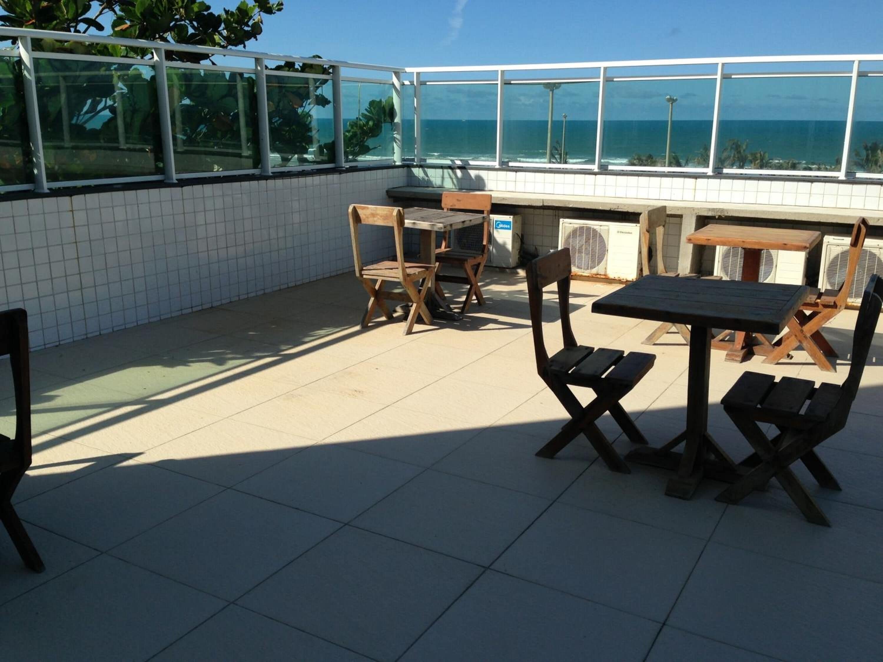 An outdoor patio with tiled flooring, wooden tables and chairs, and large windows offering a view of the ocean.
