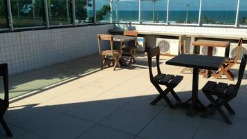 An outdoor patio with tiled flooring, wooden tables and chairs, and large windows offering a view of the ocean.
