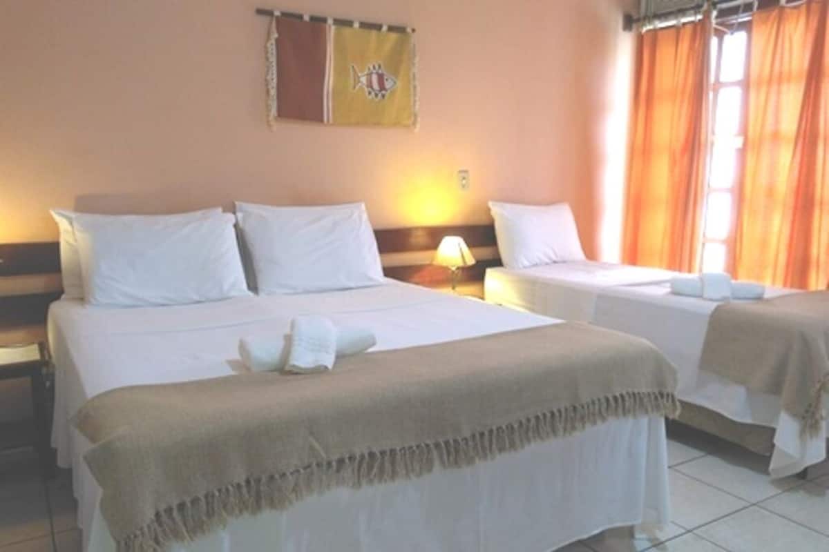 Ein Hotelzimmer mit zwei Betten, jedes mit weißer Bettwäsche und einer beige farbenen Decke. Über den Betten hängt eine Wandflagge, und zur Rechten ist ein Fenster mit orangefarbenen Vorhängen zu sehen.