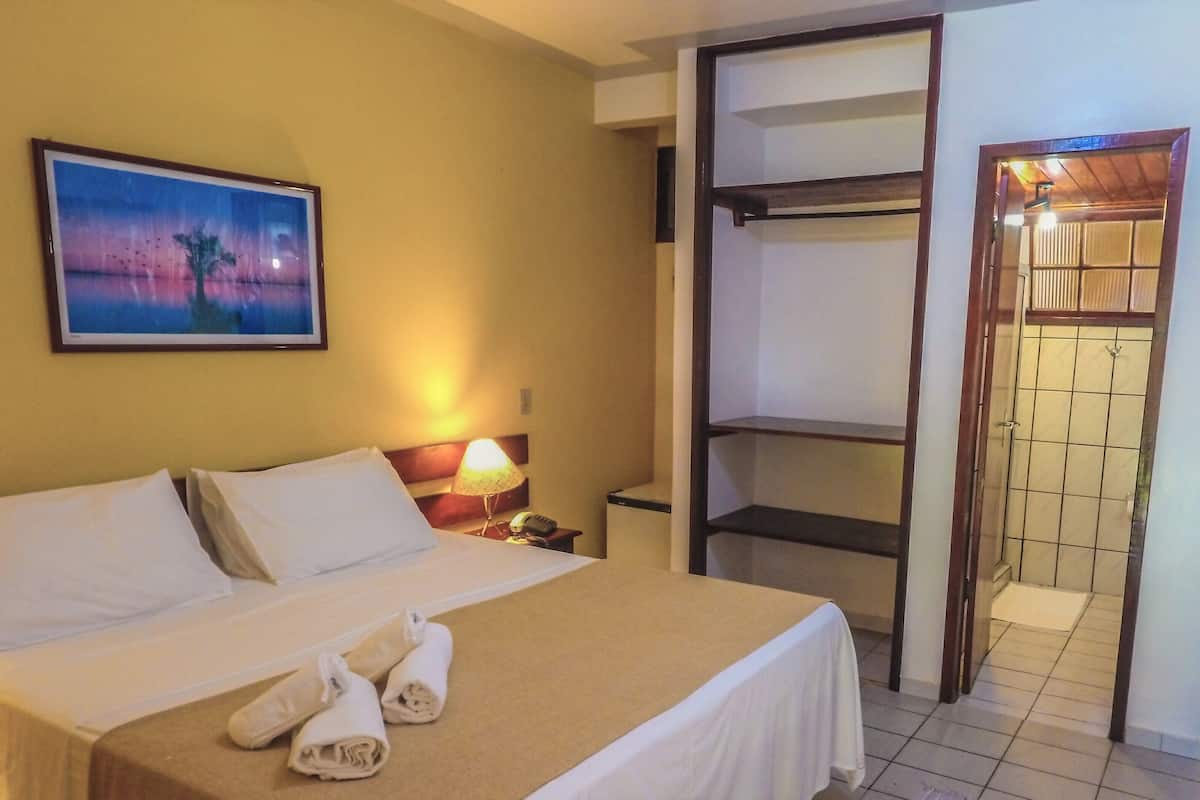 Ein Hotelzimmer mit Bett, Nachttisch, Lampe, Gemälde, Fernseher und einem Badezimmer mit Fliesenwand.