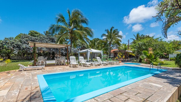 A rectangular swimming pool with a stone deck, surrounded by white lounge chairs and a covered seating area. - Pousada Aldeia Portuguesa (Santa Cruz Cabralia)