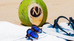 A beach scene with a coconut drink, sunglasses, and flip-flops on a towel. - Nord Easy Ondas do Atlântico (João Pessoa)