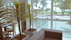 A modern interior with a sofa, chairs, and potted plants near large windows overlooking a pool and palm trees. - Nord Easy Ondas do Atlântico (João Pessoa)
