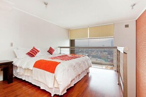 Un dormitorio con una cama grande, una mesita de noche y vistas a un paisaje urbano desde un balcón.