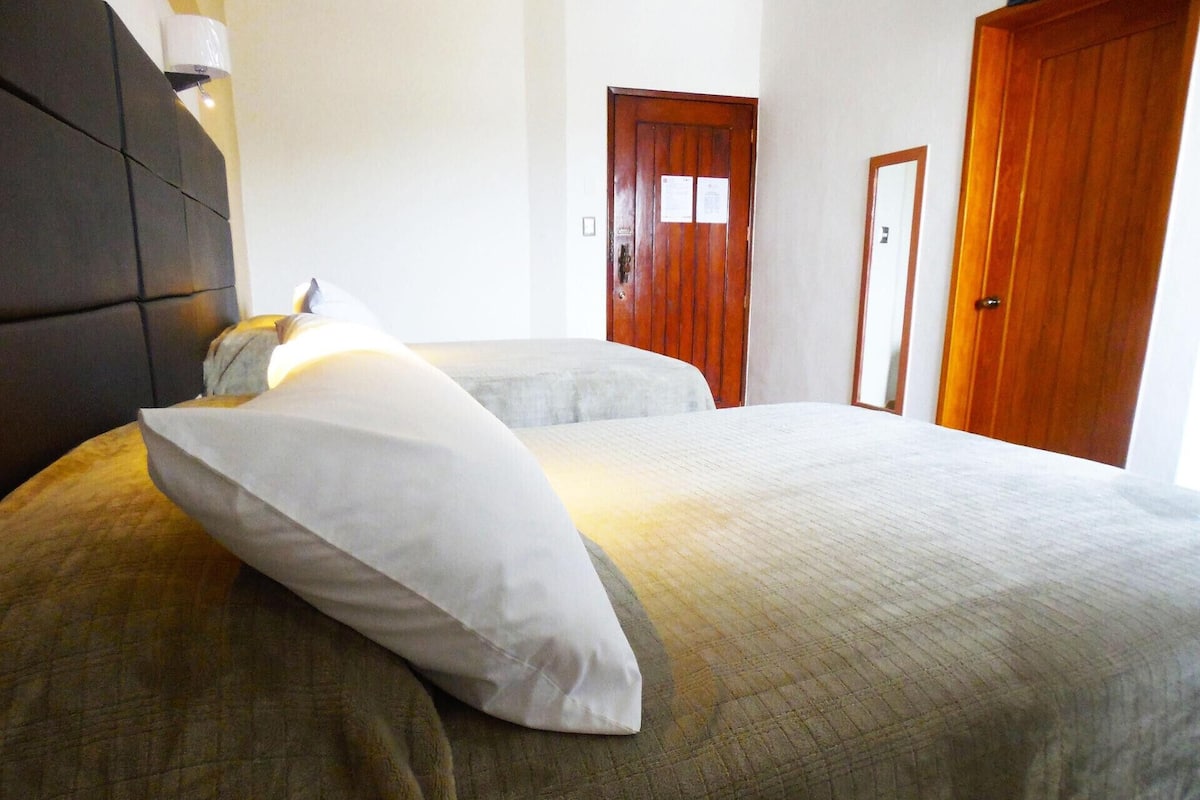 A hotel room with a large bed, a bedside table, a wall-mounted lamp, and a wooden door.