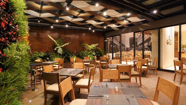 A restaurant with wooden tables and chairs, a green wall with plants, and a cityscape view through large windows. - Mercure Bogotá Calle 100 (Bogotá)
