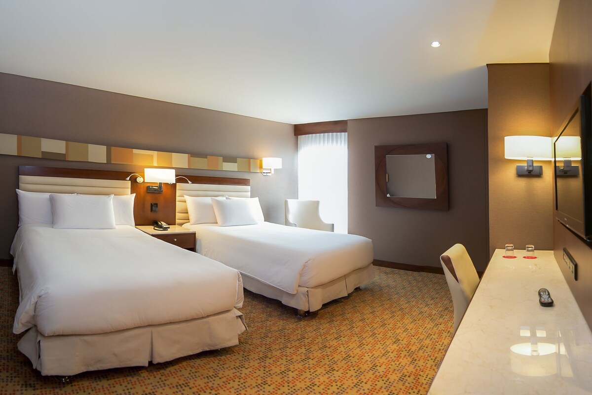 A hotel room with two beds, a desk, a chair, a TV, and a bathroom mirror.