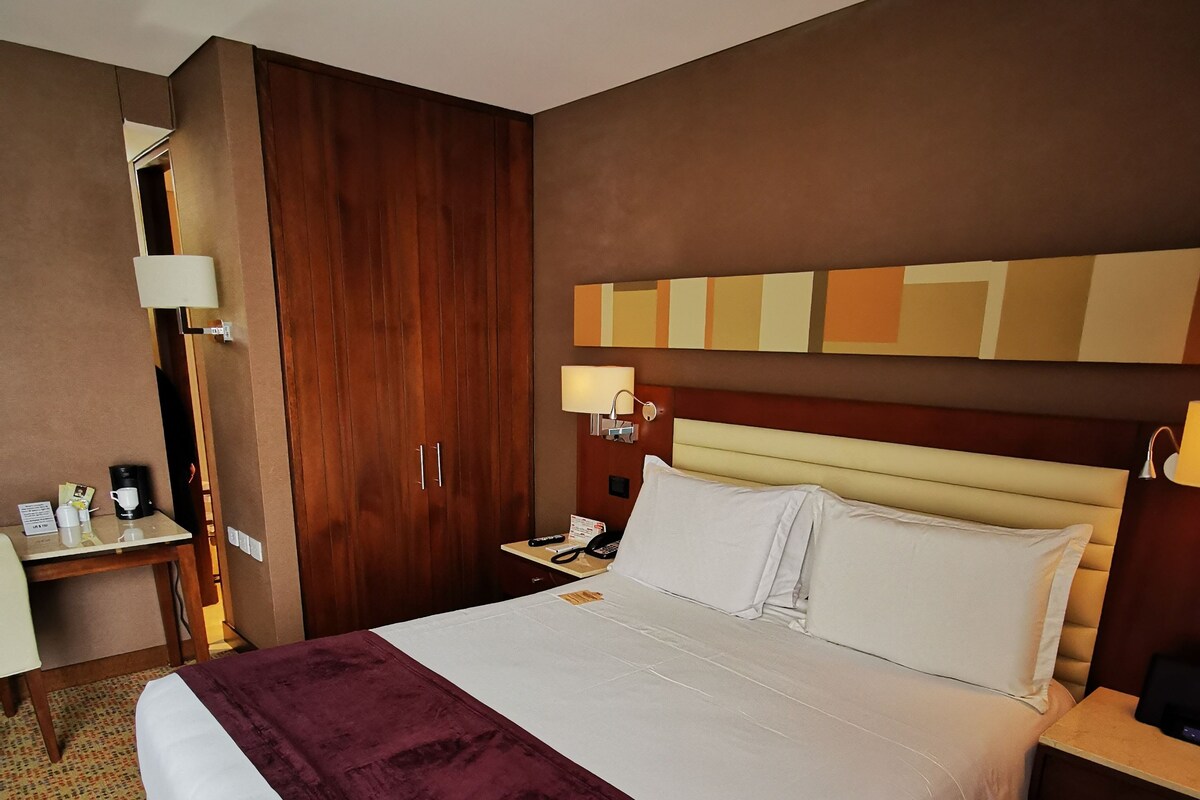 A hotel room with a large bed, a desk, a chair, a wardrobe, and a nightstand with a lamp.
